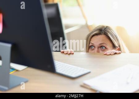 Une femme de direction effrayée cachée derrière le bureau et regardant l'écran de l'ordinateur. Banque D'Images