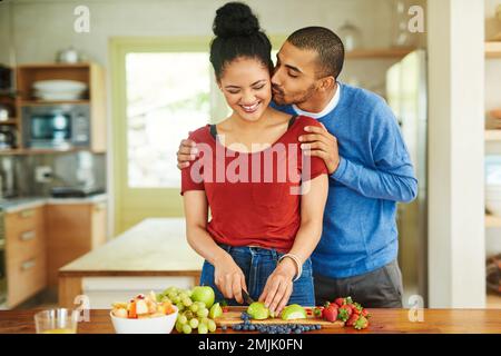 Ça a l'air bien. un beau jeune homme embrassant sa femme pendant qu'elle prépare une salade de fruits dans la cuisine. Banque D'Images