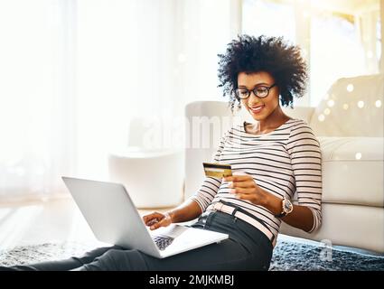 C'est la façon simple de faire les choses. une jeune femme joyeuse faisant des achats en ligne sur son ordinateur portable tout en étant assise sur le sol à la maison. Banque D'Images