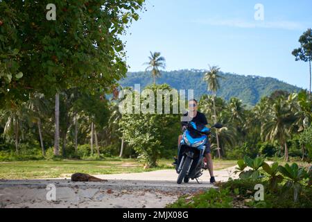 Un gars sur une moto regarde le chien dormir à l'ombre d'un arbre sur la route. Banque D'Images