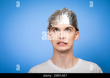 Portrait d'un jeune homme anxieux portant un chapeau en feuille d'étain et regardant dans la panique. Théories du complot et concept de paranoïa. Studio tourné sur fond bleu Banque D'Images