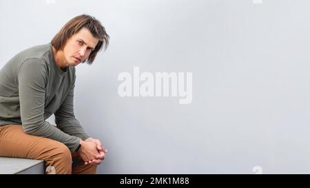 Homme millénaire aux cheveux longs et au look tendu près du mur gris. Un homme sérieux dans des vêtements décontractés aux tons de la terre est à la tête dans l'appareil photo. Copier l'espace. Banque D'Images