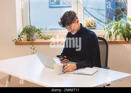 Un entrepreneur aux cheveux bruns avec un sourire sur son visage répond à ses clients sur son téléphone mobile dans son bureau avec une lumière du jour abondante. À partir de Banque D'Images