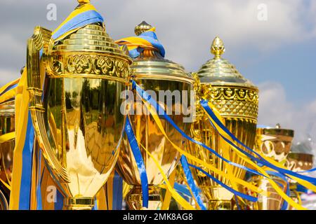 Coupes du gagnant d'or avec rubans jaunes et bleus, préparées pour les prix dans une rangée sur la table sur fond de ciel. Banque D'Images