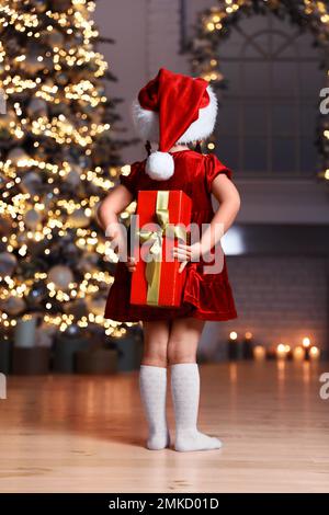 Mignon petit enfant portant chapeau de père Noël cache cadeau de Noël derrière elle dos à la maison Banque D'Images