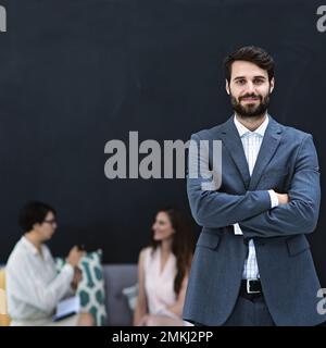 Jeune et orienté vers les objectifs. Portrait d'un jeune homme d'affaires debout dans un bureau moderne avec des collègues en arrière-plan. Banque D'Images