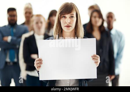 Soutenue par son équipe. Portrait d'une jeune femme d'affaires tenant un écriteau vierge avec ses collègues en arrière-plan. Banque D'Images