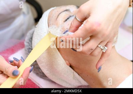 Une esthéticienne enlève un masque hydratant sur le visage d'une femme avec une spatule, procédures de cosmétologie. Banque D'Images