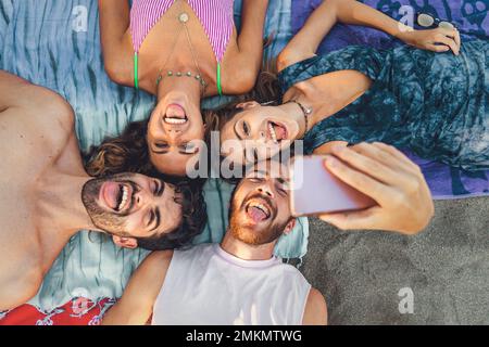 Quatre jeunes adultes qui profitent d'une journée à la plage, allongé sur des serviettes et prenant un selfie de groupe. Riant et faisant des visages stupides, ils capturent la joie et moi Banque D'Images