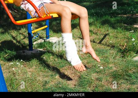 Les jambes des enfants sont bandées en plâtre - pieds nus sur une balançoire. Équipement traumatique, blessure d'enfant, ecchymoses, entorse, fracture Banque D'Images