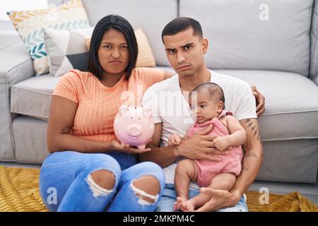 Jeune couple hispanique avec bébé tenant la banque de porc sceptique et nerveux, frowning bouleversé à cause du problème. personne négative. Banque D'Images