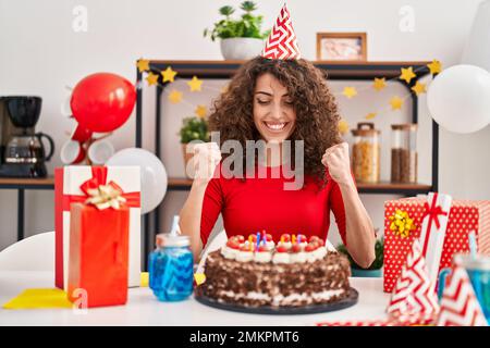 Femme hispanique aux cheveux bouclés célébrant l'anniversaire tenant le grand gâteau de chocolat criant fier, célébrant la victoire et le succès très excité avec le rais Banque D'Images