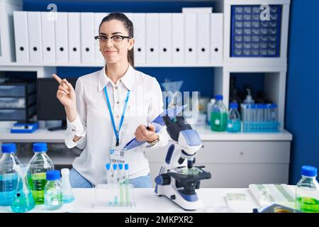 Jeune femme brunette travaillant au laboratoire scientifique pointant du doigt vers le côté montrant une publicité, sérieux et calme visage Banque D'Images