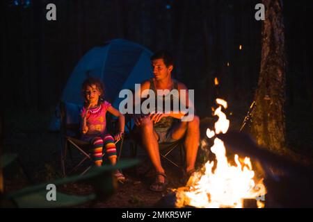 Papa et fille s'assoient la nuit près du feu en plein air en été dans la nature. Voyage de camping en famille, rassemblements autour du feu de camp. Fête des pères, Banque D'Images