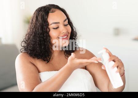 Femme noire surdimensionnée tenant un flacon et versant une lotion hydratante pour le corps à portée de main, en utilisant un nouveau produit de soin de la peau après le bain Banque D'Images