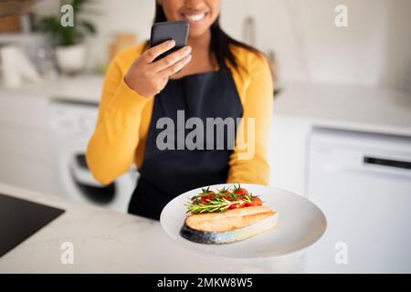 Joyeuse femme afro-américaine millénaire en tablier prenant la photo d'un plat de poisson sur un smartphone Banque D'Images