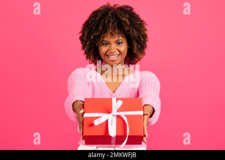 Gai joli jeune noir curly femme dans une ambiance décontractée avec bretelles donne boîte rouge avec cadeau Banque D'Images