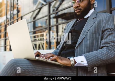 Un homme d'affaires noir en costume utilise un ordinateur portable à l'extérieur tout en étant assis sur un banc. Gros plan Banque D'Images