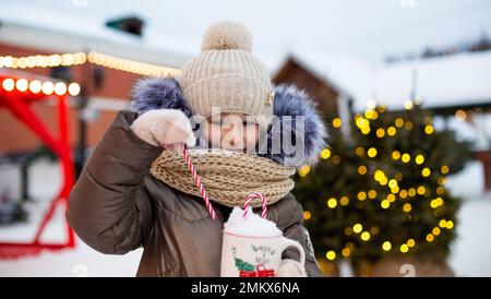 Fille avec mug avec neige, canne à sucre et inscription Merry et Bright dans ses mains en plein air dans des vêtements chauds en hiver sur le marché de fête. Guirlande lumineuse ga Banque D'Images