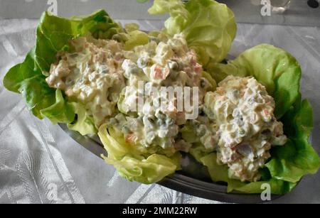 Délicieuse salade russe traditionnelle avec carottes, pommes de terre, petits pois, jambon, œufs et concombres mélangés à de la mayonnaise dans de la laitue sur une table. Banque D'Images