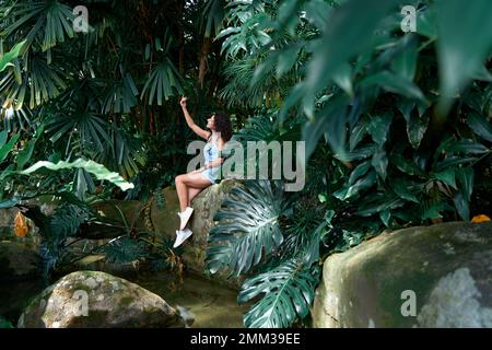 Une belle fille pose sur des rochers près de l'eau dans la jungle parmi les plantes. Banque D'Images