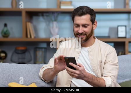 Gros plan. Un jeune homme beau dans une chemise beige est assis à la maison sur le canapé, utilise un téléphone. Banque D'Images