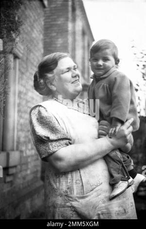 1950s, historique, une dame portant une robe chasuble sur une robe tenant un petit garçon, son petit-fils, à l'extérieur d'une maison, Angleterre, Royaume-Uni. Banque D'Images