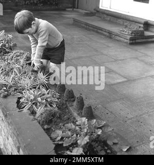 1960s, historique, un jeune garçon, en short et un pull à col roulé, jouant seul à l'extérieur sur un jardin patio, faisant de petits châteaux de sable de terre! Banque D'Images