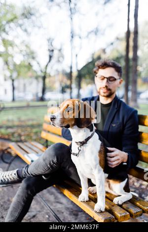 un jeune homme élégant est assis avec son chien de beagle sur un banc de parc Banque D'Images