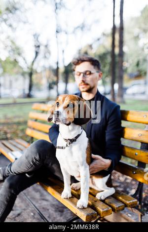 un jeune homme élégant est assis avec son chien de beagle sur un banc de parc Banque D'Images