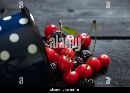 cerises et mûres dans une tasse sur une table en bois noir parsemée, fond de fruits, jus de fruits Banque D'Images