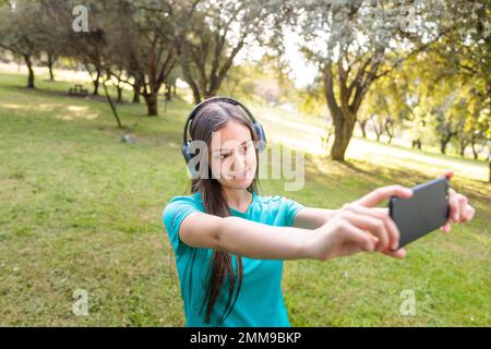 Adolescente souriante portant un t-shirt turquoise et un casque, emportant un selfie avec son mobile dans un espace naturel Banque D'Images