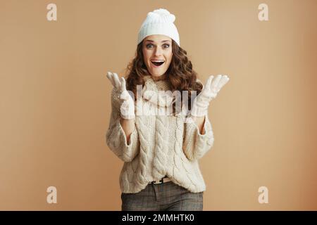 Bonjour hiver. Portrait d'une femme élégante surprise de 40 ans en chandail beige, mitaines et chapeau isolés sur fond beige. Banque D'Images