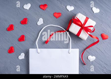 Concept d'achat en ligne. Sac à provisions blanc brillant et cadeau avec noeuds en ruban rouge sur fond gris Ultimate tendance avec coeurs rouges et blancs. Valenti Banque D'Images