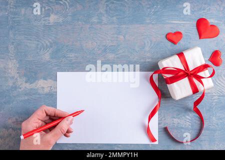 Main féminine tient un stylo rouge sur une feuille de papier blanc vierge, à côté d'un cadeau avec des coeurs rouges sur un fond bleu grunge en bois, vue du dessus, espace de copie Banque D'Images