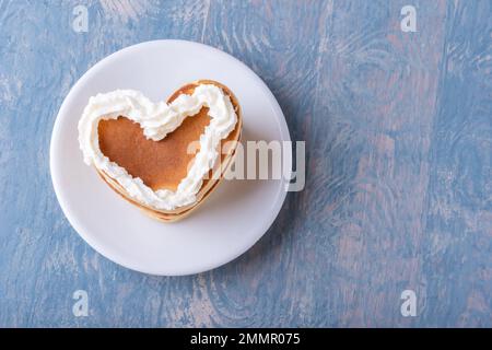 Petit déjeuner créatif pour la Saint-Valentin. Petit déjeuner romantique au lit. Crêpe en forme de coeur maison décorée de crème blanche sur une assiette blanche sur un woo bleu Banque D'Images