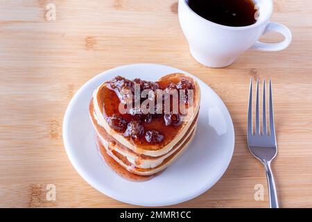 Petit déjeuner créatif au lit. Crêpes faites maison en forme de coeur avec confiture de baies sur une assiette blanche, fourchette et tasse avec café ou cacao sur un fond en bois. P Banque D'Images