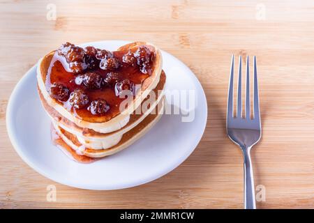 Petit déjeuner créatif au lit. Crêpes faites maison en forme de coeur avec confiture de baies sur une assiette blanche et une fourchette sur un fond en bois. Carte postale pour la Saint-Valentin Banque D'Images