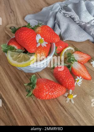 Vue de fraises entières et coupées en deux à l'intérieur d'un bol blanc et sur une table en bois, décorée de tranches de citron et de pétales de fleurs, tissu décoratif Banque D'Images