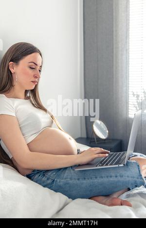 Belle femme enceinte se trouve sur le lit et utilise un ordinateur portable. Banque D'Images