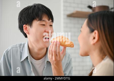 Joyeux et amusant jeune ami asiatique nourrissant un croissant à sa petite amie dans la cuisine, ayant du temps amusant sur le petit déjeuner dans la cuisine. Banque D'Images