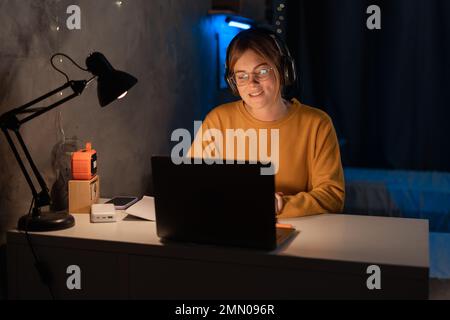 Une étudiante étudie en ligne, parcourt le concept d'e-learning. Jeune fille concentrée écoutant le tuteur d'Internet tard dans la nuit dans le dortoir utilisant un ordinateur portable Banque D'Images