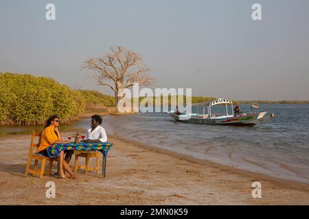 Sénégal, delta de Saloum classé au patrimoine mondial de l'UNESCO, couple assis et buvant un cocktail de fruits sur la plage d'une île couverte de mangroves devant un baobab et un pirogue traversant la rivière Banque D'Images