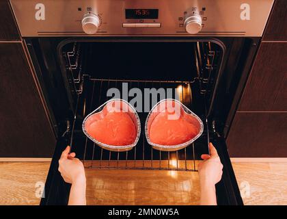 Une femme met deux moules en aluminium avec de la pâte rouge dans le four pour un gâteau en forme de cœur pour la Saint-Valentin. Banque D'Images