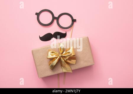 Vue à plat du cadeau de Père emballé avec moustache et lunettes sur fond rose, beaucoup d'espace de copie. Maquette de carte de vœux amusante. Banque D'Images