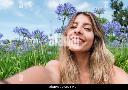 Portrait selfie de la belle jeune Latina, femme colombienne aux cheveux longs, assise dans le parc avec des fleurs derrière elle, elle est heureuse souriante et looki Banque D'Images