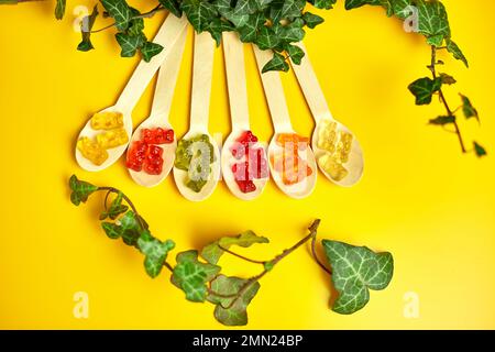 Composition à plat avec des ours en gelée de légumes sains colorés dans une cuillère en bois écologique avec des feuilles vertes sur fond jaune, des vitamines, des bonbons sains, à Banque D'Images