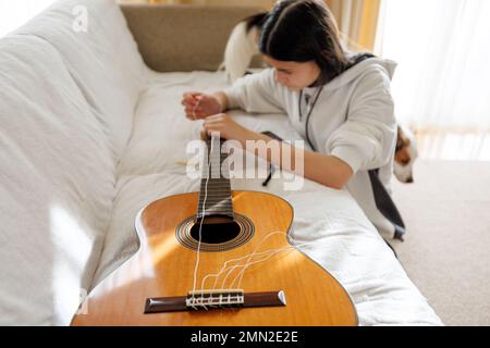 Adolescente avec de longs cheveux bruns et des cordes à capuchon blanches changeant dans la guitare acoustique classique tout en étant assis sur le sol. Chien en arrière-plan. Banque D'Images