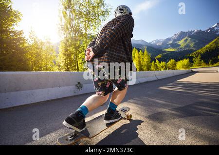 Man Skater en casque sur la route de montagne à sa longue planche près du sommet de la neige au matin d'été Kazakhstan Banque D'Images