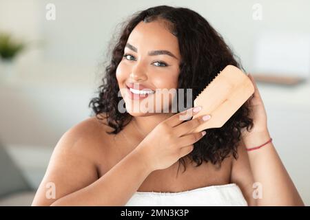 Portrait du corps heureux positive dame se brossant ses cheveux maussaux sombres et souriant à l'appareil photo, intérieur de la maison Banque D'Images